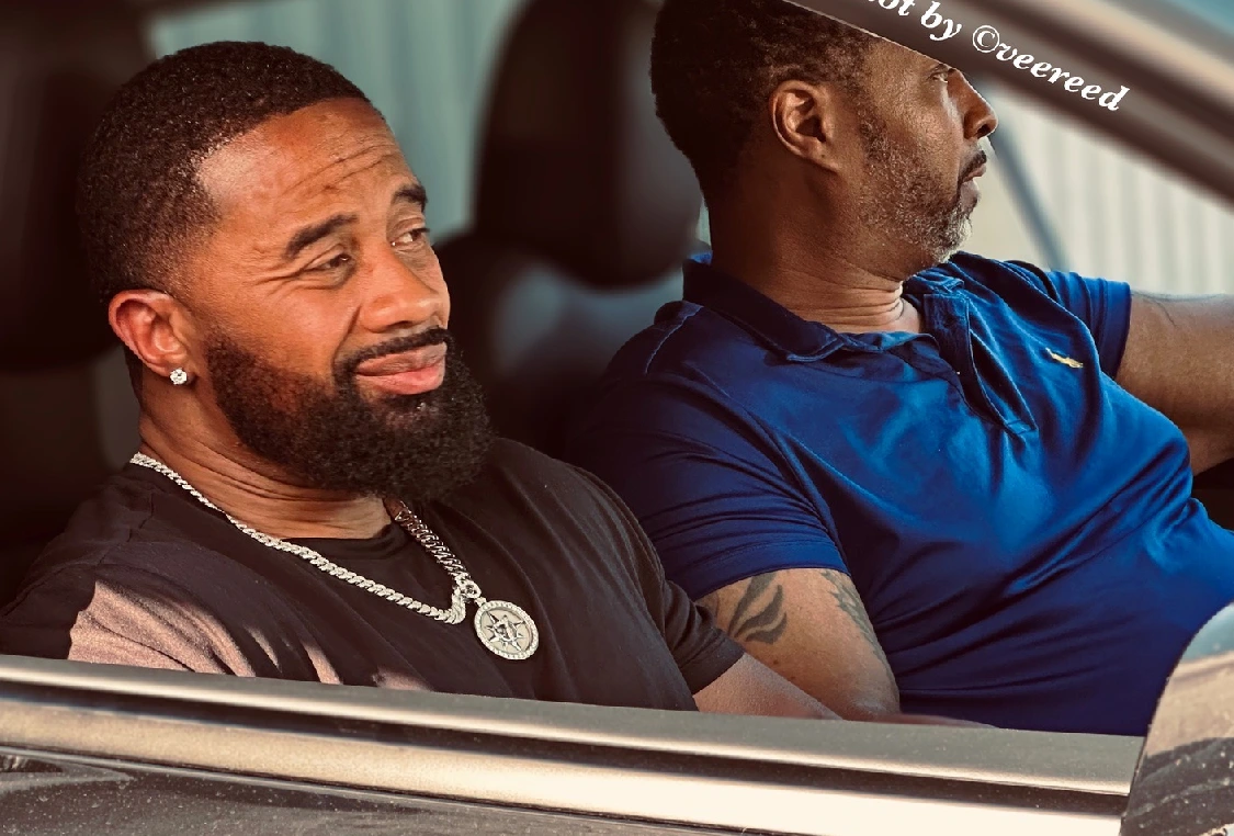 Two men sitting in a car with one man wearing a necklace.