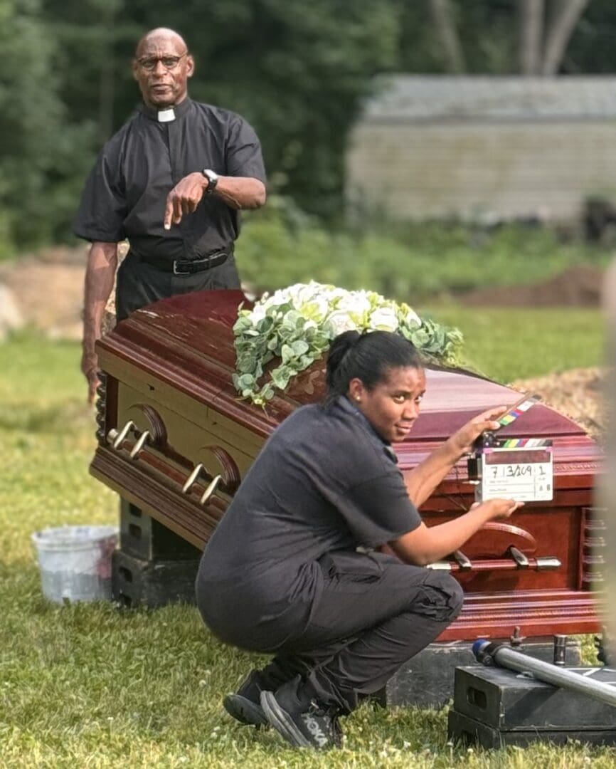 Two men in black shirts and one is kneeling next to a casket.